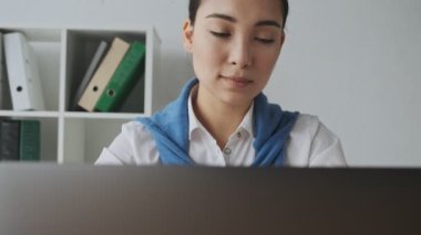 Asyalı genç bir sekreter, ofiste çalışırken kameraya gülümseyerek bakmaktansa dizüstü bilgisayarını kullanıyor.