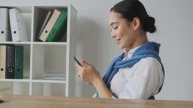 Çekici bir Asyalı sekreter kadın ofiste otururken cep telefonunu kullanıyor.