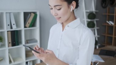 Mutlu gülümseyen, genç, Asyalı, kablosuz kulaklıklı sekreter kadın ofiste otururken cep telefonunu kullanıyor.