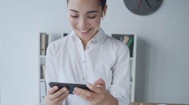 Kablosuz kulaklıklı çekici Asyalı genç bir iş kadını cep telefonundan komik bir şey izliyor ve ofiste otururken gülüyor.