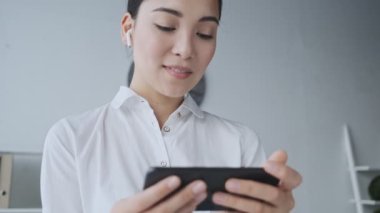 Kablosuz kulaklıklı mutlu Asyalı genç bir iş kadını ofiste otururken cep telefonundan güzel bir şey izliyor.