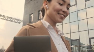Modern ofis binalarının yanında yürüyen, kulaklık takan Asyalı genç bir iş kadınının alt görüşü.