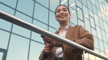 Kulaklık takan kendinden emin Asyalı genç bir iş kadını modern ofis binalarının yanında duruyor.