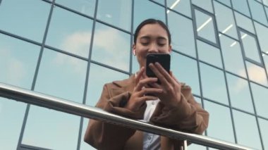 Kulaklık takan çekici Asyalı genç bir iş kadını modern ofis binalarının yanında akıllı telefonunu kullanıyor.