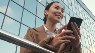 Kulaklık takmış güzel Asyalı genç bir iş kadınının en alt görüntüsü modern ofis binalarının yanında duran akıllı telefonunu kullanıyor.