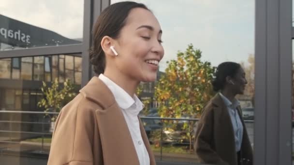 現代的なオフィスビルの近くを歩いていると 笑顔の若いアジアの女性がイヤホンを使って電話で話しています — ストック動画