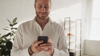 Gülümseyen genç bir adam akıllı telefonuyla daktilo yazıyor. Evdeki oturma odasında duruyor.