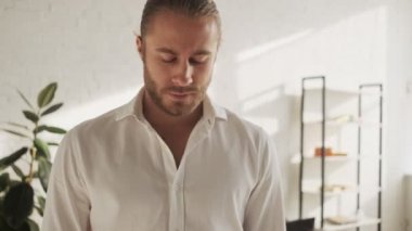 Kendinden emin genç bir adam evdeki oturma odasında dururken kameraya bakıyor.