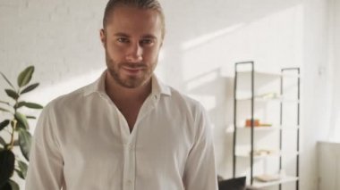 Yakışıklı bir genç adam evdeki oturma odasında dururken kameraya bakıyor.