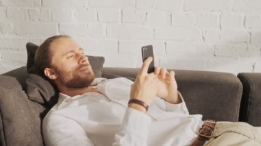 Sakin bir genç adam evdeki oturma odasındaki kanepede yatarken akıllı telefonunu kullanıyor.
