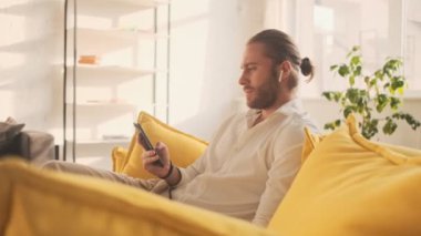 Telsiz kulaklıklı sakin bir genç adam evdeki oturma odasındaki kanepede otururken akıllı telefonunu kullanıyor.