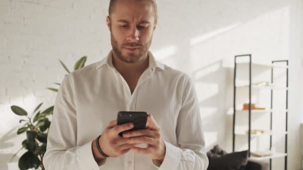 Atrakcyjny Młody Mężczyzna Wykonuje Gest Zwycięzcy Trzymając Smartfona Salonie Domu — Wideo stockowe