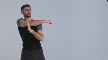 Spor kıyafetleri içinde ciddi şekilde konsantre olmuş genç bir sporcu ısınma egzersizleri yapıyor. Stüdyodaki gri arka plan üzerinde kollarını esnetiyor.