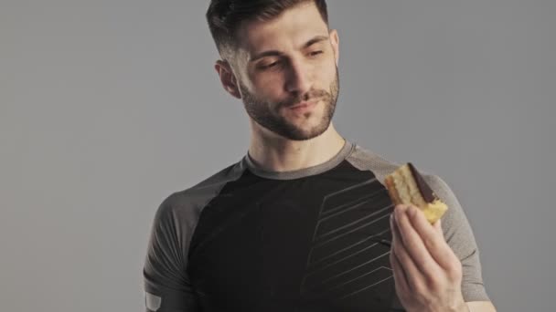 スポーツウェアのハンサムな若い男は彼の腕の中でケーキを保持しており スタジオでグレーの背景に隔離されたストップジェスチャーを示しています — ストック動画