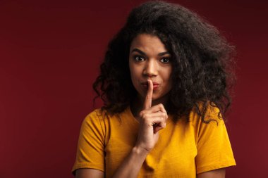 Koyu kırmızı arka plan üzerinde izole edilmiş poz veren sevimli Afrikalı genç bir kadının fotoğrafı sessizlik hareketi gösteriyor..