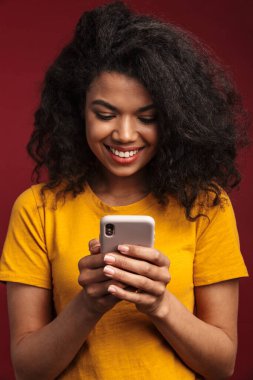 Kırmızı arka planda izole edilmiş, kıvırcık saçlı, gülümseyen ve cep telefonu kullanan güzel esmer Afro-Amerikalı kadın.