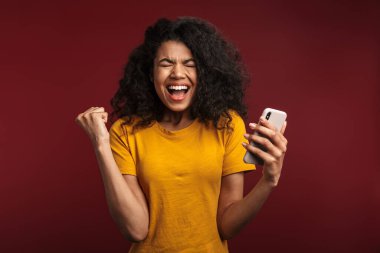 Kırmızı arka planda izole edilmiş, kıvırcık saçlı ve cep telefonu kullanan güzel esmer Afrikalı Amerikalı kadın.