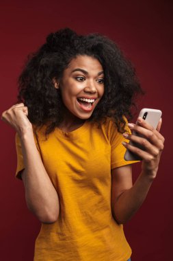 Kırmızı arka planda izole edilmiş, kıvırcık saçlı ve cep telefonu kullanan güzel esmer Afrikalı Amerikalı kadın.