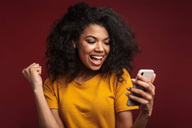 Kırmızı arka planda izole edilmiş, kıvırcık saçlı ve cep telefonu kullanan güzel esmer Afrikalı Amerikalı kadın.