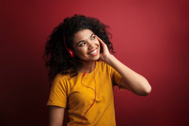 Mutlu, pozitif, iyimser bir Afrikalı kadının, koyu kırmızı duvar arkasında izole bir şekilde poz verip kulaklıkla müzik dinlediği bir fotoğraf..