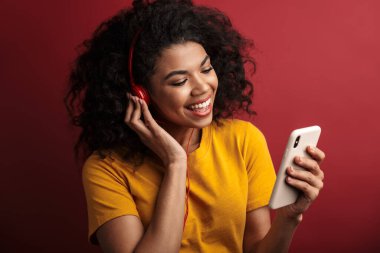 Güzel, esmer, Afro-Amerikalı, kulaklıklı, gülümseyen ve cep telefonunu kırmızı arka planda saklanmış bir kadın.