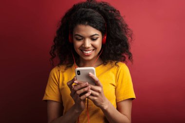 Güzel, esmer, Afro-Amerikalı, kulaklıklı, gülümseyen ve cep telefonunu kırmızı arka planda saklanmış bir kadın.