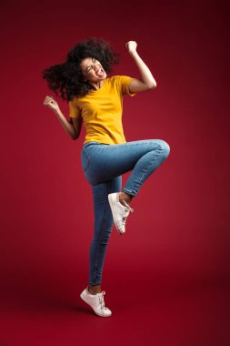 Güzel esmer, kıvırcık saçlı, kızıl arka planda dans edip eğlenen Afro-Amerikan kadınının tam boy görüntüsü.
