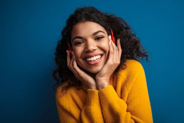Güzel, genç ve mutlu bir Afrikalı kadının görüntüsü. Mavi duvar arkasında izole bir şekilde poz veriyor. Kulaklıkla müzik dinliyor..