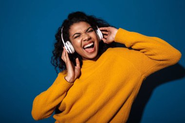 Heyecanlı, genç bir Afrikalı kadının görüntüsü. Mavi duvar arkasında izole bir şekilde duruyor. Kulaklıkla müzik dinliyor..