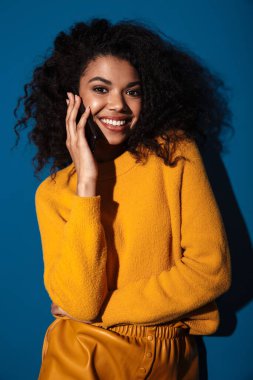 Oldukça neşeli gülümseyen Afrikalı bir kadının görüntüsü. Mavi duvar arkasında izole edilmiş poz veriyor. Cep telefonuyla konuşuyor..