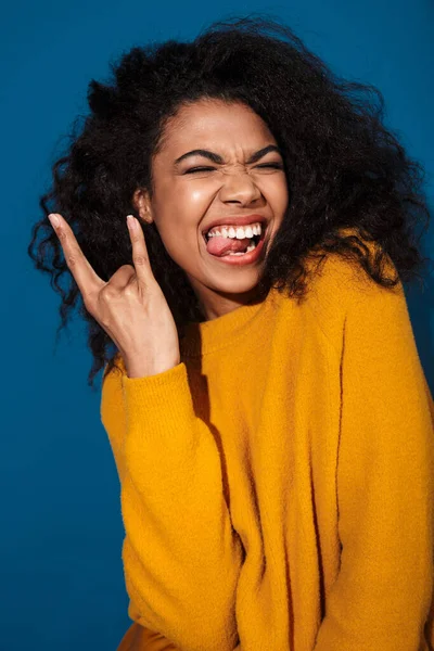 Mavi duvar arkasında izole bir şekilde poz veren güzel, duygusal, genç bir Afrikalı kadın görüntüsü kaya jesti yapıyor..