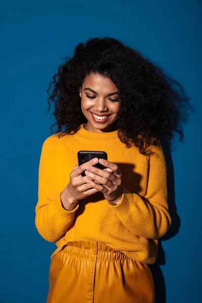 携帯電話を使って青い壁の背景に隔離されたアフリカの女性の笑顔のかなり肯定的なイメージ — ストック写真