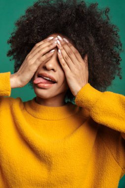Yeşil arka planda tek başına duran, yüzünü ekşiten ve elleriyle yüzünü kapatan gülümseyen Afrikalı genç bir kadının portresi.