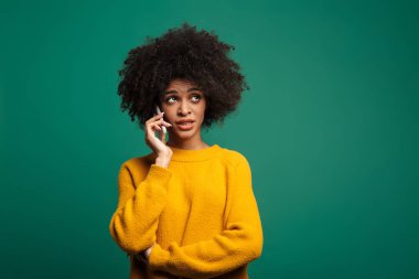Kafası karışmış genç bir Afrikalı kadının portresi yeşil arka planda tek başına dikilip cep telefonuyla konuşuyor.