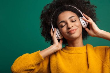 Koyu mavi duvar arkasında izole edilmiş, kulaklıkla müzik dinleyen memnun bir Afrikalı kıvırcık kadın görüntüsü..