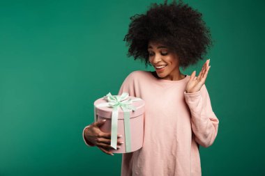 Yeşil arka planda tek başına duran, elinde büyük bir hediye kutusu olan, eşofman üstü giyen gülümseyen Afrikalı genç bir kadının portresi.