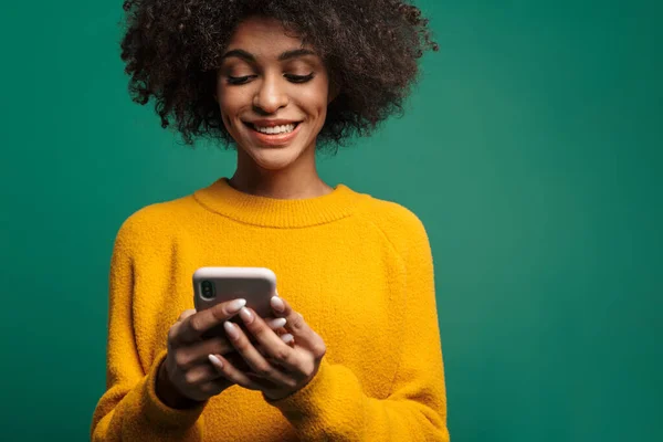 携帯電話を使って緑の背景に隔離されたセーターを着た若いアフリカ人女性の肖像画 — ストック写真