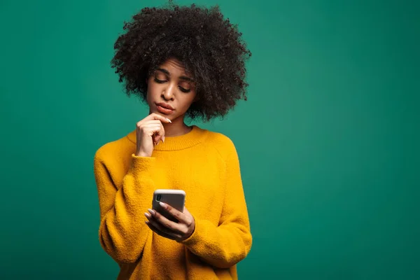 携帯電話を使って緑の背景に隔離されたセーターを着た混乱した若いアフリカ人女性の肖像画 — ストック写真