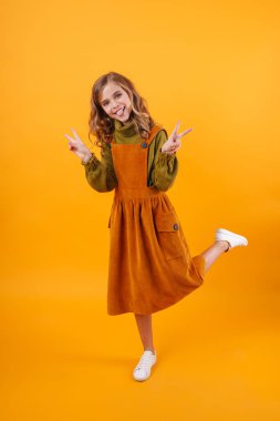 Mavi arka planda izole bir şekilde duran, barış jesti gösteren, gülümseyen küçük bir kız çocuğu.