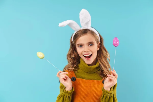 照片上的金发碧眼的小女孩在玩具兔子耳朵里笑着 抱着蓝色背景下的彩蛋 — 图库照片