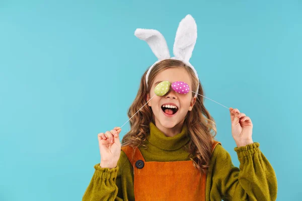 照片中快乐的金发女孩在玩具兔子耳朵里与蓝色背景的彩蛋嬉戏 — 图库照片