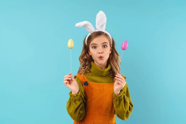 照片上 金发碧眼的小女孩站在玩具兔子的耳朵里 抱着彩蛋 把亲吻的嘴唇与蓝色背景隔离在一起 — 图库照片