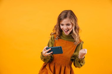 Sarı duvar arkasında izole edilmiş, mutlu, tatlı bir kız çocuğunun görüntüsü cep telefonuyla oyunlar oynayarak kazanan hareketi yap.