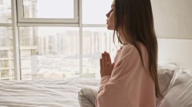 Sakin, genç ve çekici bir kız meditasyon yapıyor. Yatakta oturuyor ve evde dua eden bir pozisyonda el ele tutuşuyor.
