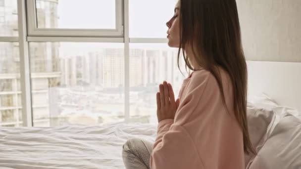 穏やかな若い魅力的な女の子は瞑想をしていますベッドの中に座って自宅で祈りのポーズで手を握って — ストック動画