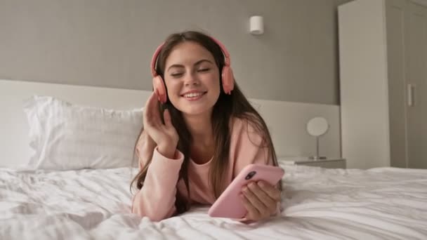 一个漂亮的小女孩正躺在床上和家里用她的智能手机和耳机听音乐 — 图库视频影像