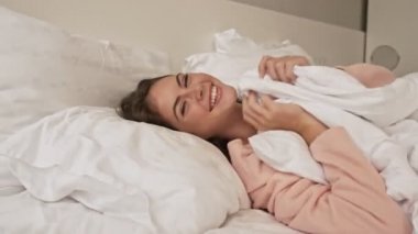 Gülümseyen genç bir kız eğleniyor ve gülüyor. Evdeki yatakta yatıyor.