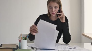Sarışın, güzel bir iş kadını beyaz bir ofiste kağıt belgelerle çalışırken telefonda konuşuyor.