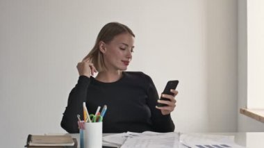 Çekici sarışın bir iş kadını, beyaz bir ofiste çalışırken akıllı telefonuna bakıyor.