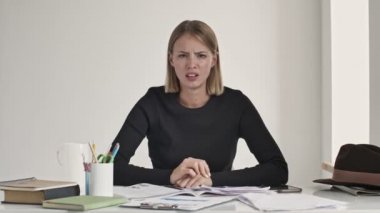 Hoşnutsuz genç sarışın bir kadın beyaz bir ofisteki masada otururken baş parmak hareketi yapıyor.